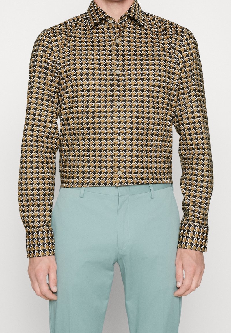 Ein Hemd mit Knopfleiste im braun-schwarzen Hahnentrittmuster, mit klassischem Kragen und langen Ärmeln. Kombiniert mit hellblauen Hosen.