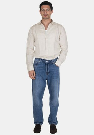 Uomo in piedi con una mano in tasca, che indossa una camicia beige chiaro abbottonata, jeans blu e scarpe scure su sfondo bianco semplice.