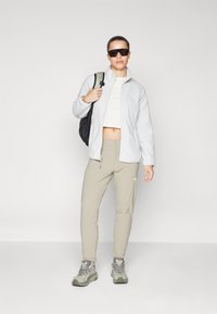 Giacca in felpa grigia chiara con zip, pantaloni beige e sneakers grigie. Il modello porta uno zaino nero e indossa occhiali da sole scuri.