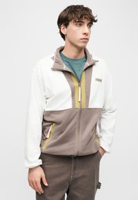 Νέος άνδρας που φοράει λευκό και ταουπέ φλις μπουφάν με φερμουάρ και πράσινες και κίτρινες λεπτομέρειες, στέκεται με το ένα χέρι στην τσέπη, κοιτάζοντας προς τα δεξιά.