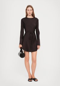 Svart langermet kjole med livbelte, glatt stoff, rund halslinning og asymmetrisk kant. Modellen holder en rund svart håndveske og bruker flate sandaler.