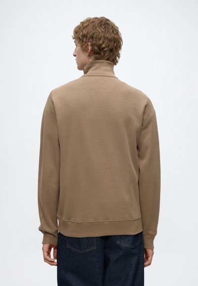Personne aux cheveux bouclés portant un pull à col roulé beige et un jean bleu foncé, de dos sur un fond clair uni.