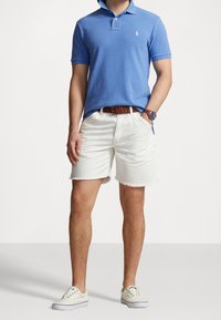 Polo blu con colletto e un piccolo logo, abbinato a pantaloni corti di jeans bianchi con orli sfilacciati. Indossato con Sneakers chiare e una cintura marrone.