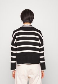 Osoba z krótkimi ciemnymi włosami, ubrana w sweter w czarno-białe paski i białe spodnie, stojąca na tle jasnym, jednolitym.