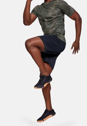 Homme portant une chemise verte à motifs et un short noir, levant le genou haut, chaussé de baskets noires à semelles beige, sur fond blanc.