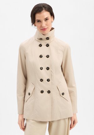 Frau trägt eine beige zweireihige Jacke mit schwarzen Knöpfen und hohem Kragen, steht vor einem schlichten weißen Hintergrund.