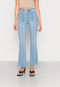 Högmidjade utsvängda jeans i ljusblå denim med kontrastsömmar och rå fåll. Kombineras med svarta klacksandaler med öppen tå.