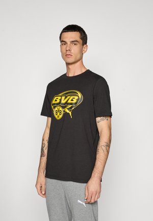 Mand med kort mørkt hår iført en sort BVB-logo t-shirt og grå Puma sweatpants, stående foran en ensfarvet lys baggrund.