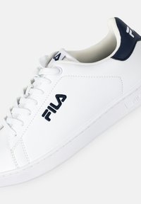Sneaker bianco con tomaia in pelle texturizzata, dettagli blu navy sul tallone e logo ricamato in evidenza. Presenta un tradizionale sistema di allacciatura.