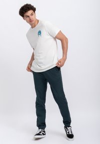 T-shirt bianca con una grafica colorata sul petto, abbinata a pantaloni jogger verde scuro e sneakers bianco e nere.