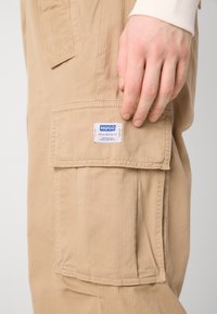 Beige cargo pants with a textured fabric, featuring a large side pocket and a logo tag with blue text. Seam detailing is visible.