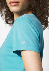 Pánské tričko s krátkým rukávem v tyrkysové barvě vyrobené z bavlny, s růžovou vyšitou nášivkou "Polo" na levém rukávu. Hladká textura a uvolněný střih.