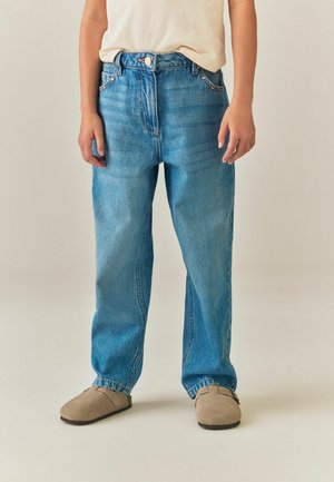 Person wearing light blue straight-leg jeans, beige slip-on shoes, and a tucked-in white shirt, standing on a plain background.