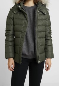 Doudoune vert olive avec capuche doublée de fausse fourrure, design matelassé, fermeture éclair et deux poches latérales. Portée par-dessus un pull gris.