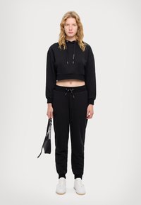 Felpa nera corta con cappuccio e cordini, abbinata a pantaloni neri. Il modello tiene una piccola borsa nera, indossando sneaker bianche.