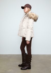 Giubbotto puffo beige con cappuccio in pelliccia sintetica, abbinato a leggins marroni e stivali di pelle marrone al ginocchio con fibbie.