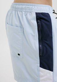 Lichtblauwe zwemshorts met donkerblauwe accenten, een elastische tailleband en een ritse zak. Gemaakt van lichtgewicht, waterbestendig materiaal.