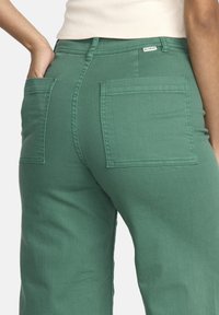 Pantaloni in misto cotone verde con vita alta, due tasche posteriori quadrate e dettagli cuciti, caratterizzati da una texture liscia e una vestibilità casual.