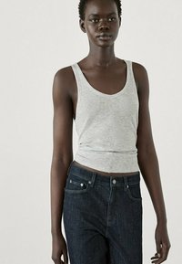Femme portant un débardeur côtelé sans manches gris clair et un jean taille haute bleu foncé, debout devant un fond uni clair.