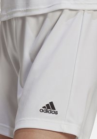 Białe szorty sportowe wykonane z gładkiego materiału, z haftowanym czarnym logo Adidas na dolnej lewej stronie.