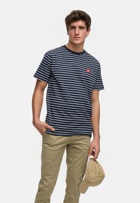 Marine-stripet t-skjorte med hvite linjer, korte ermer, og en liten rød logo på brystet. Kombinert med beige bukser og holder en lys brun caps.