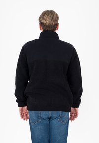 Giacca in pile nera con colletto alto e superficie testurizzata, con un pannello nero liscio sulla parte superiore della schiena. Indossata con jeans blu.