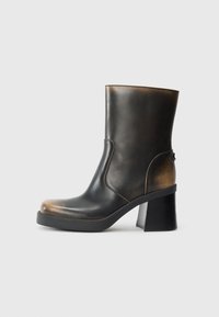 Bottine en cuir noir avec une finition brillante, bout rond, accents en tan contrastés, un talon carré et un détail de fermeture éclair sur le côté.