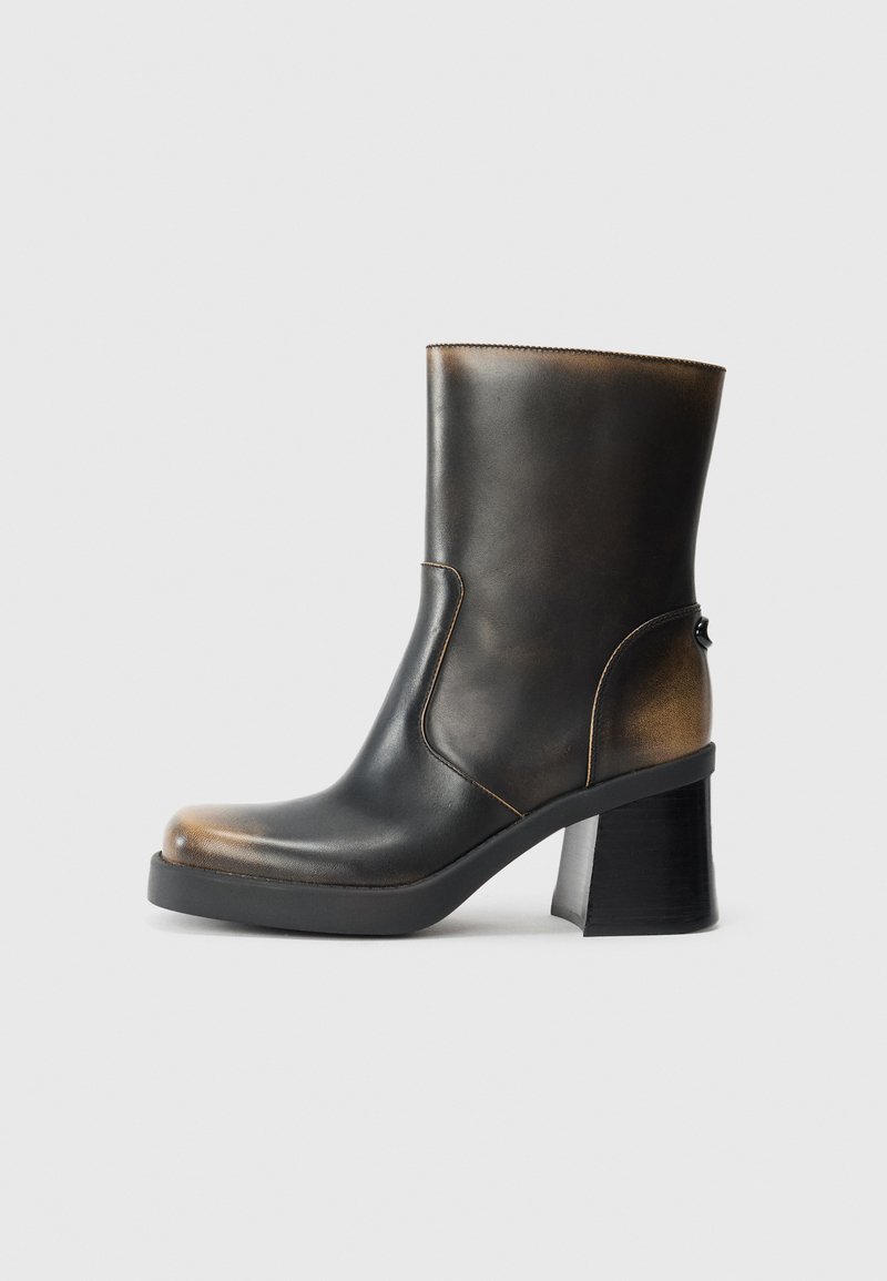 Bottine en cuir noir avec une finition brillante, bout rond, accents en tan contrastés, un talon carré et un détail de fermeture éclair sur le côté.