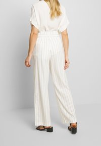 Bredbyxlade, högmidjade byxor i vitt tyg med vertikala ljusgrå ränder. Slät textur, minimalistisk design, parat med svarta klacksandaler.