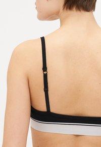 Fekete bralette állítható pántokkal; az alján kontrasztos szürke sávval és sima, puha szövettextúrával rendelkezik.