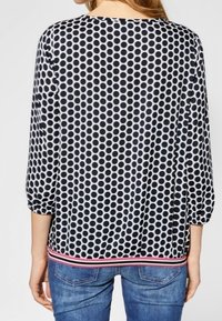 Kvinna iklädd en blus med svartvita prickar och trekvartsärmar, med randig nederkant, i kombination med blå denimjeans, sedd bakifrån.