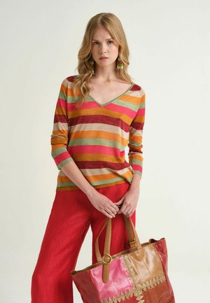 Frau mit langen blonden Haaren trägt einen mehrfarbig gestreiften Pullover und rote Hosen, hält eine große Patchwork-Ledertragetasche mit gewebten Details.