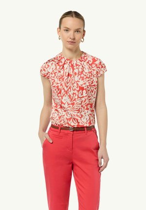 Floral-patterned, short-sleeve top in coral and white, featuring pleats at the neckline. Paired with tailored red trousers and a brown belt.