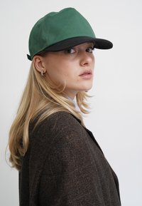 Grüner Hut mit schwarzem Schirm, aus Canvas gefertigt. Das Modell trägt einen braunen strukturierten Blazer und schaut leicht nach rechts.