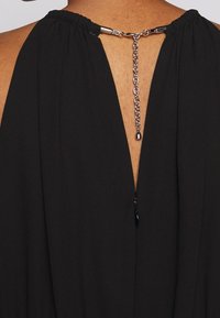 Vue de dos d'un vêtement noir avec un décolleté en V profond maintenu par une chaîne argentée et un fermoir à la nuque.