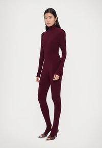Bordo spalvos ilgomis rankovėmis vientisas kūno apdaras su apykaklės imitacija, aptempiantis siluetą, lygios tekstūros ir pailgintos rankovės, dengiančios rankas.