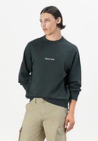 Sudadera verde oscuro con cuello redondo acanalado, mangas largas y texto bordado en blanco en el pecho; combinada con pantalones cargo de color beige claro.
