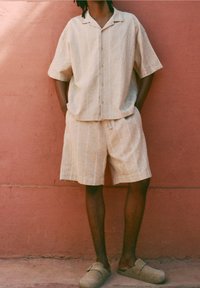 Lichtbeige linnen overhemd met korte mouwen en knopen, gecombineerd met bijpassende shorts. De outfit heeft subtiele strepen en een ontspannen pasvorm.
