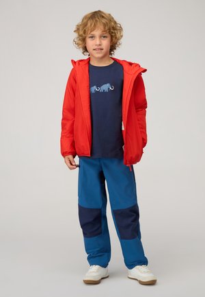 Rote Zip-Jacke, blaues T-Shirt mit Elefanten-Grafik, blaue Hose mit dunkelblaue Patches, weiße Schuhe, lockige Haare.
