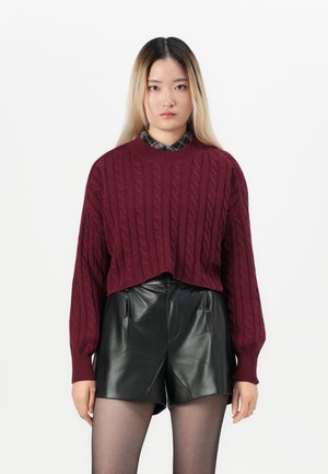 Pull en tricot torsadé bordeaux à manches longues, design court associé à un short en cuir noir et des collants noirs transparents.