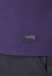 Camiseta morada hecha de un tejido suave, con una etiqueta metálica plateada que lleva la marca "Dropside" cerca del dobladillo. Diseño simple y sin adornos.