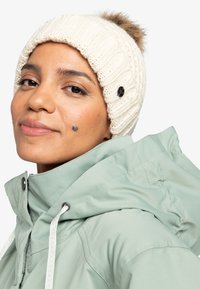 Strikket crème-farvet hue med et tekstureret mønster og en pelskvast på toppen, båret med en lysegrøn jakke med høj krave og justerbare snore.