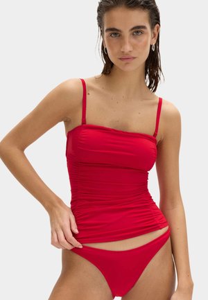 Kobieta z mokrymi włosami, ubrana w czerwony marszczony top tankini i pasujące bikini, pozująca na jasnym, jednolitym tle.