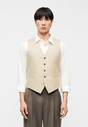 Man draagt een beige gilet met knopen over een wit overhemd met opgerolde mouwen en losse bruine broek, staand tegen een effen achtergrond.