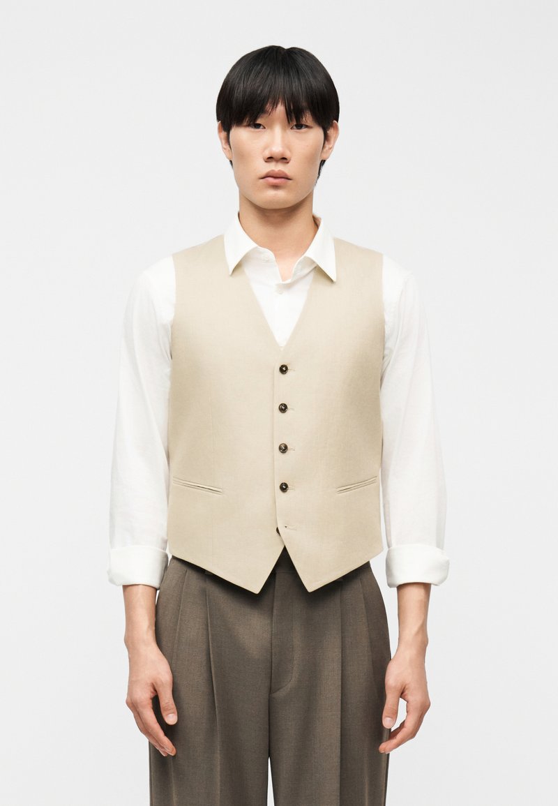 Άνδρας που φοράει μπεζ γιλέκο με κουμπιά πάνω από λευκό πουκάμισο με ανασηκωμένα μανίκια και χαλαρό καφέ παντελόνι, στέκεται μπροστά σε απλό φόντο.