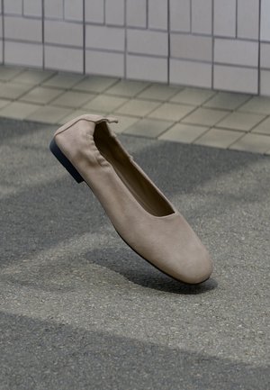 Beiger Flachschuh mit rundem Zehenbereich und elastischem Hinterteil, schwebt über einer gefliesten und gepflasterten Bodenfläche.