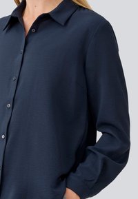 Camicia a maniche lunghe blu navy realizzata in tessuto liscio, con collo a camicia, bottoni frontali e una vestibilità rilassata con maniche a costura.