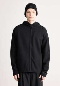 Μαύρο φούτερ με φερμουάρ, υψηλό γιακά και τσέπες καγκουρό. Κατασκευασμένο από απαλό ύφασμα, διαθέτει μινιμαλιστικό σχέδιο και ελαστικά μανίκια. Συνδυάζεται με σκουφάκι.