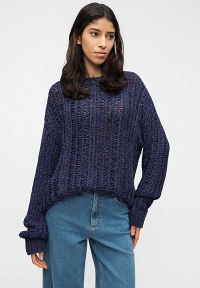Pull en maille bleu marine avec un design lâche et ajouré, rayures texturées et manches larges, porté avec un jean bleu clair.