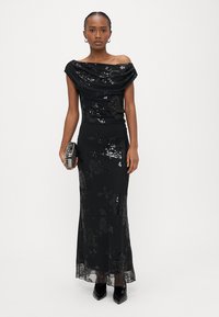 Sieviete, kas valkā melnu ziedu raksta cilpiņu vakarkleitu ar atsegtiem pleciem, turot melnu nelielu rokassomiņu, stāvot pret vienkāršu baltu fonu.